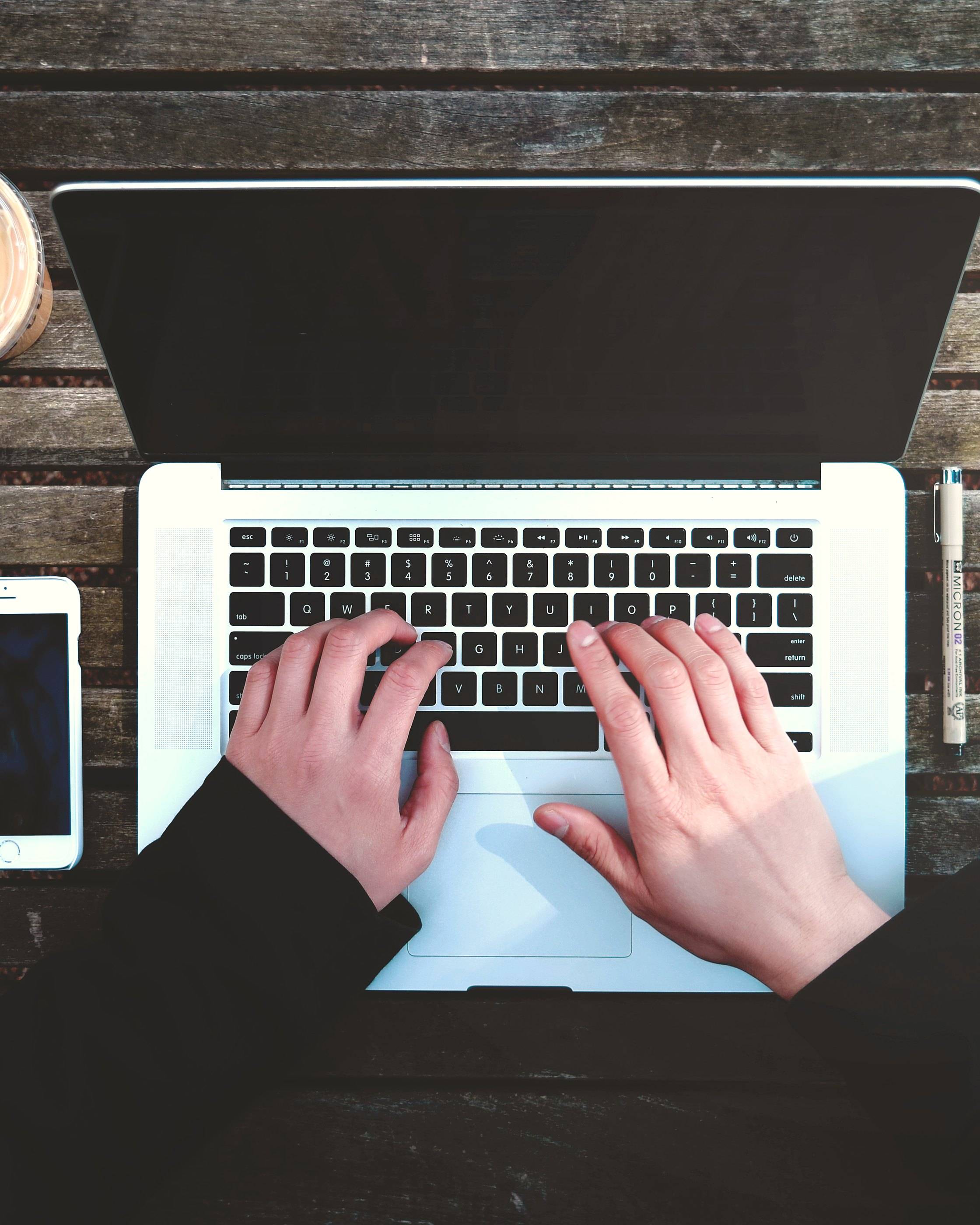 Hands typing on a laptop on a dark wooden table, a phone and a set square beside it