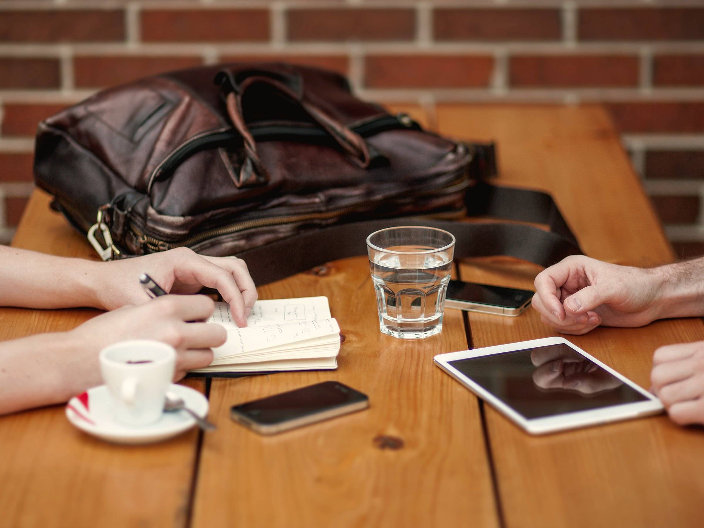 Two people at a wooden café table with a notebook of page sketches, two phones, a tablet and an espresso